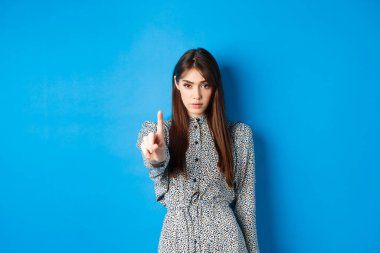 Ciddi bir kadın kendinden emin görünüyor ve parmağını sallıyor, hayır diyor, seni durduruyor, aynı fikirde değil ve onaylamıyor, mavi arka planda giyinmiş duruyor.