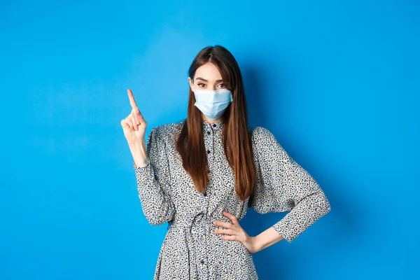 Personnes en bonne santé et concept de pandémie covid-19. Femme sérieuse en masque médical pointant vers le coin supérieur gauche, debout sur fond bleu — Photo