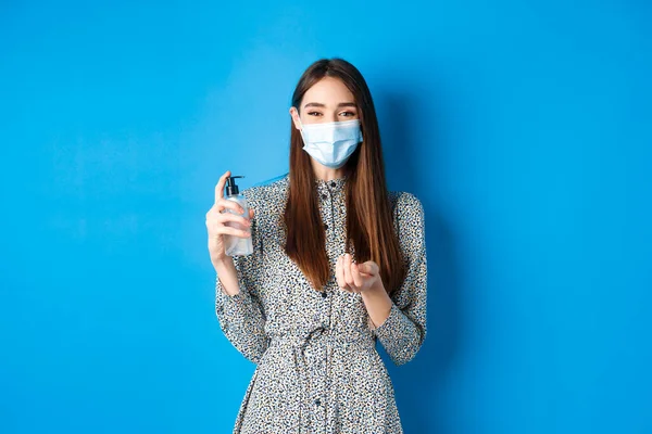 Covid-19，社会疏离和保健概念。戴着医疗面罩的漂亮女孩，手握消毒瓶，站在蓝色的背景上 — 图库照片