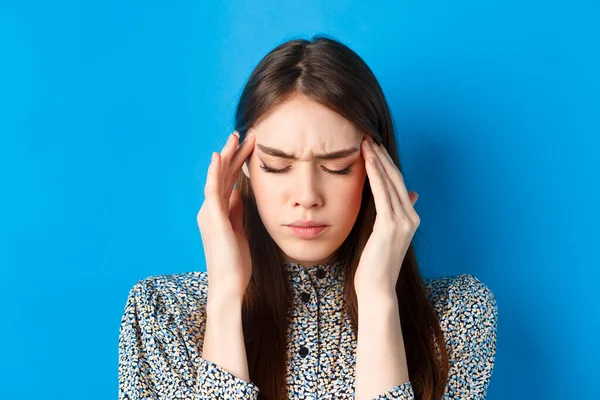 Close-up retrato de jovem se sentir doente, tocando templos da cabeça e franzindo a testa de dor de cabeça, tendo enxaqueca, de pé sobre fundo azul — Fotografia de Stock