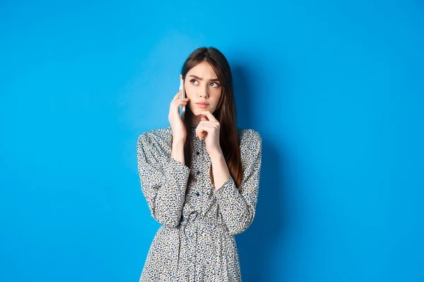 Mujer pensativa hablando por teléfono y pensando, mirando a un lado pensativo, de pie sobre fondo azul —  Fotos de Stock