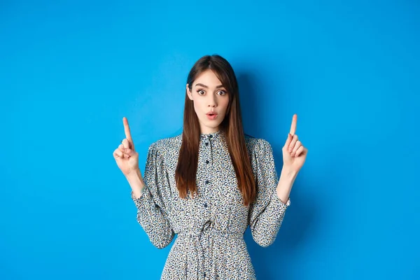 Emocionado señora bonita en vestido decir wow, mirando intrigado por la publicidad, señalando los dedos hacia arriba en el logotipo, de pie sobre el fondo azul — Foto de Stock