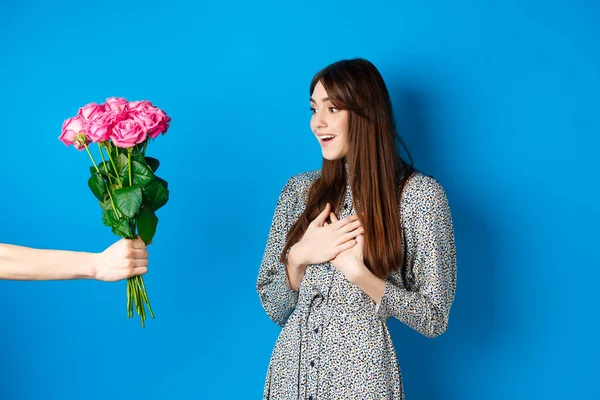 Koncept Valentýna. Obrázek překvapené krásné dívky při pohledu na ruku s kyticí květin, obdrží překvapivé růže od milence, stojí na modrém pozadí — Stock fotografie