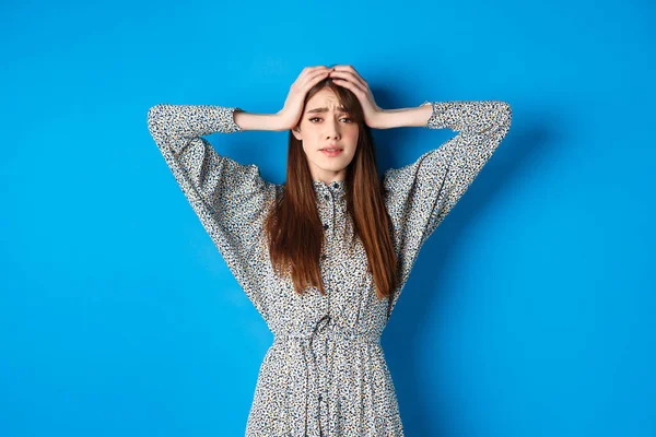 Απογοητευμένη ενήλικη γυναίκα με φυσικά μαλλιά να κρατιέται χέρι-χέρι στο κεφάλι και πανικόβλητη, να δείχνει τρομαγμένη και φοβισμένη, να στέκεται ντυμένη σε μπλε φόντο — Φωτογραφία Αρχείου