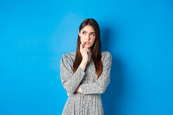 Troubled pensive meisje kijken opzij op logo, bijten vingernagel en denken, staan op blauwe achtergrond in lange jurk — Stockfoto