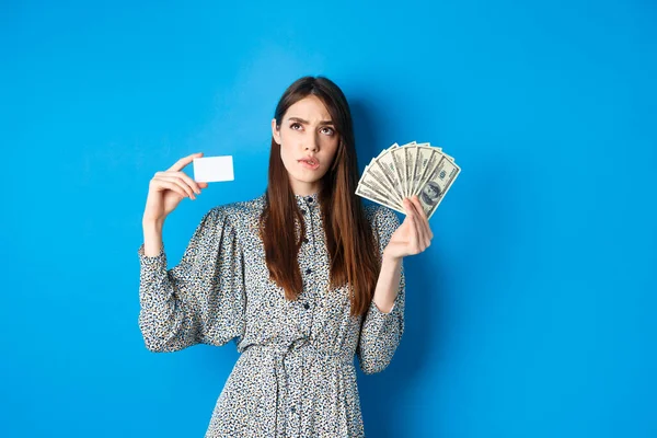 Compras. Chica reflexiva mirando hacia arriba y mordiendo el labio, contando en mente o pensando, sosteniendo la tarjeta de crédito de plástico con billetes de dólar, de pie sobre fondo azul — Foto de Stock