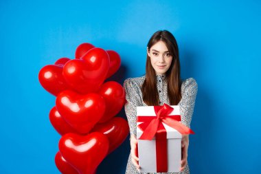 Sevgililer Günü. Romantik ve tatlı bir kız hediye kutusunu sevgilisine veriyor ve gülümsüyor, kalp balonlarının yanında duruyor, mavi arka plan.