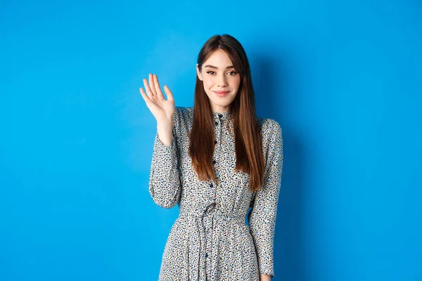 Nette lächelnde Frau winkt zur Begrüßung, grüßt mit schüchternem Grinsen und steht im Kleid vor blauem Hintergrund — Stockfoto