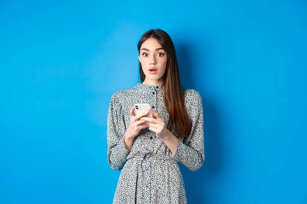 Woman look surprised after using mobile phone, standing on blue background — Stock Photo, Image