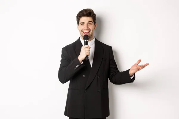 Portrait of handsome man in black suit singing a song, holding microphone and giving speech, standing against white background — Stock Photo, Image