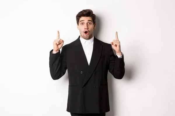 Retrato de empresario guapo sorprendido en traje negro, diciendo wow y mirando sorprendido, señalando los dedos hacia arriba en el espacio de copia, de pie sobre el fondo blanco —  Fotos de Stock