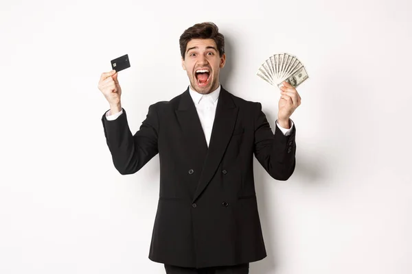 Retrato de hombre de negocios guapo en traje negro, mostrando tarjeta de crédito y dinero, gritando de alegría y emoción, de pie sobre fondo blanco — Foto de Stock
