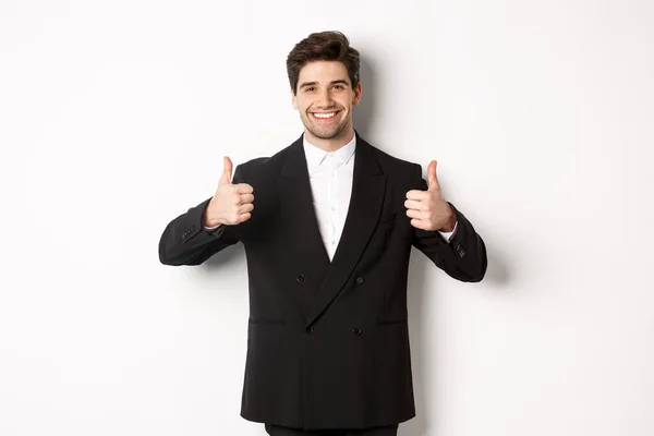 Concept of new year party, celebration and lifestyle. Image of attractive happy businessman in formal suit, showing thumbs-up and smiling, like and approve, standing over white background — Stock Photo, Image