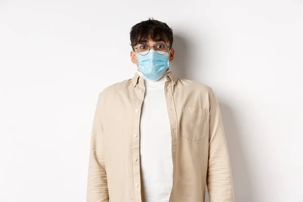 Jeune homme en masque médical stérile de covid-19 regardant surpris à la caméra, debout sur fond blanc. Coronavirus, concept de quarantaine et de distanciation sociale — Photo