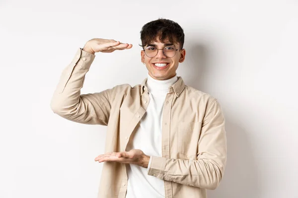 Pessoas reais. Cara moderna feliz em óculos mostrando tamanho grande, moldando caixa e sorrindo, mostrando algo grande, de pé sobre fundo branco — Fotografia de Stock