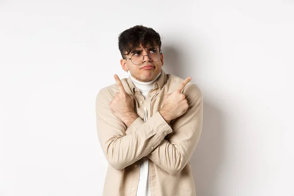 Indecisive funny young man having troubles with making choice, pointing fingers sideways and frowning perplexed, deciding between two variants, standing on white background — Stock Photo, Image