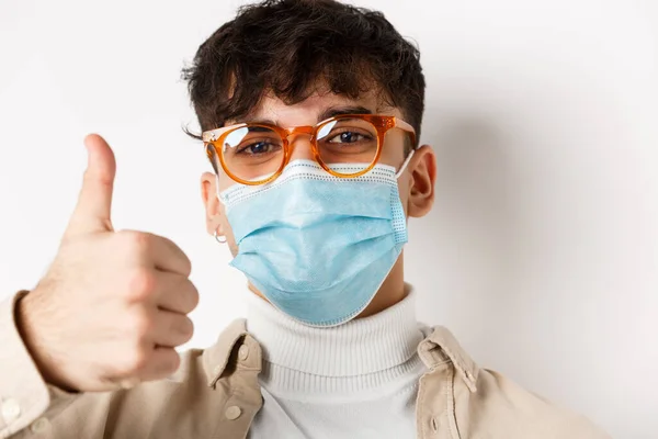 Portrait of young man in glasses and medical mask showing thumb up, smiling with eyes, standing on white background. Covid-19 and pandemic concept — Stock Photo, Image