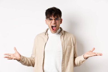 Portrait of angry frustrated man shouting and having argument, spread hands sideways and grimacing outrated, standing pissed-off on white background