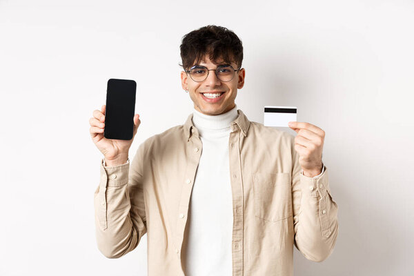 Online shopping. Natural guy in glasses showing empty smartphone screen and plastic credit card, smiling pleased, recommending bank, standing on white background