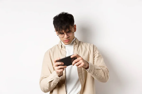 Портрет красивого молодого человека, играющего в видеоигры на мобильном телефоне, наклон смартфона, чтобы играть в гонки, стоя на белом фоне — стоковое фото