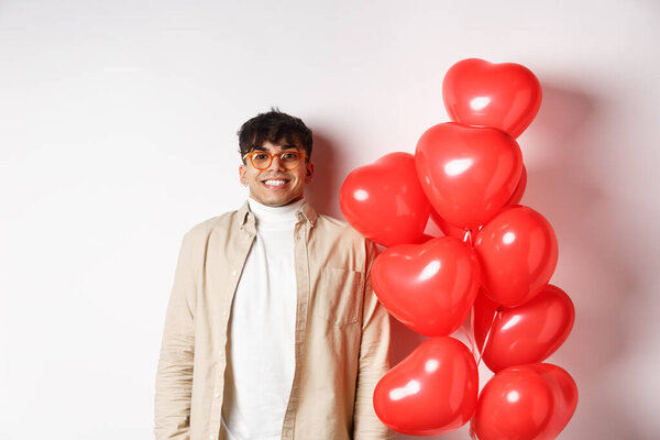 Valentines day. Excited young man smiling, looking hopeful, standing near big red hearts balloons, waiting for true love on lovers date, white background