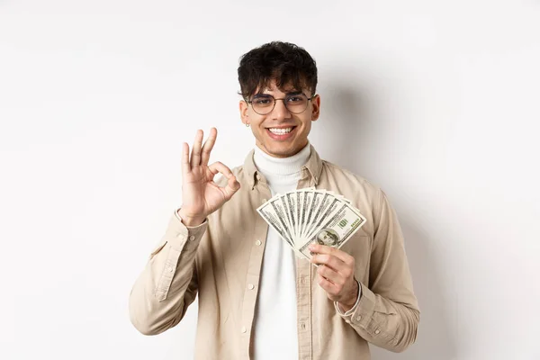 Hombre moderno guapo en gafas mostrando billetes de dólar y buen gesto, haciendo dinero, de pie con dinero en efectivo sobre fondo blanco — Foto de Stock