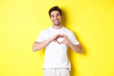 Mutlu romantik adam kalp işareti gösteriyor, gülümsüyor ve sevgisini ifade ediyor, sarı arka planda duruyor.