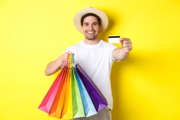 Joven sonriente comprando cosas con tarjeta de crédito, sosteniendo bolsas de compras y luciendo feliz, de pie sobre fondo amarillo —  Fotos de Stock