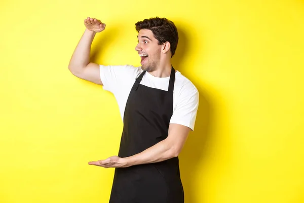 Homem olhando surpreso com algo grande, de pé em avental preto contra fundo amarelo — Fotografia de Stock