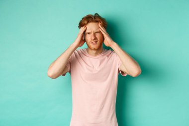 Genç kızıl saçlı bir adam kafaya dokunuyor ve başı dönüyor, baş ağrısı ya da migren hissediyor, naneli bir arka planda tişört giyiyor.