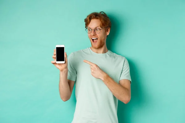 Mira esto. Guapo pelirrojo en gafas apuntando con el dedo a la pantalla del teléfono inteligente en blanco, mostrando promoción en línea, de pie sorprendido sobre fondo turquesa — Foto de Stock
