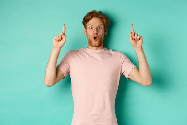 Impresionado chico guapo con el pelo rojo señalando los dedos hacia arriba, demostrar oferta promocional, de pie en camiseta sobre fondo de menta —  Fotos de Stock