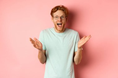 Kızıl saçlı, gözlüklü, harika haberler veren neşeli genç sakallı bir adam. Ellerinizi yana açın ve pembe arka planda sizi tebrik edin.
