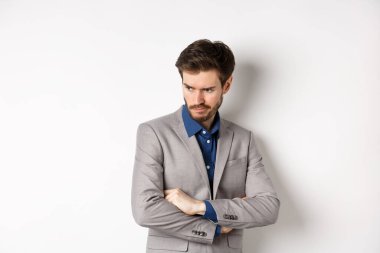 Huysuz takım elbiseli adam, adil olmayan yüzüyle yana bakıyor, göğsünde çapraz kollar ve somurtkan sinirli, beyaz arka plana karşı üzgün duruyor.