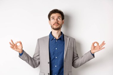 Takım elbiseli sakin yönetici meditasyonla rahatlıyor, yukarı bakıyor ve Zen Mudra işaretinde el ele tutuşuyor, beyaz arka planda duruyor.