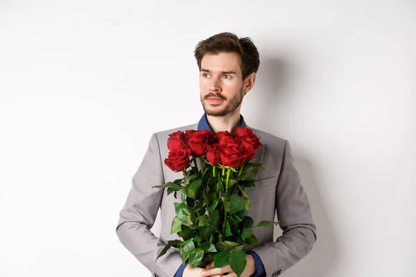 Schöner Freund im Anzug bei einem romantischen Date, hält einen Strauß roter Rosen in der Hand und schaut nachdenklich nach links, vor weißem Hintergrund stehend — Stockfoto