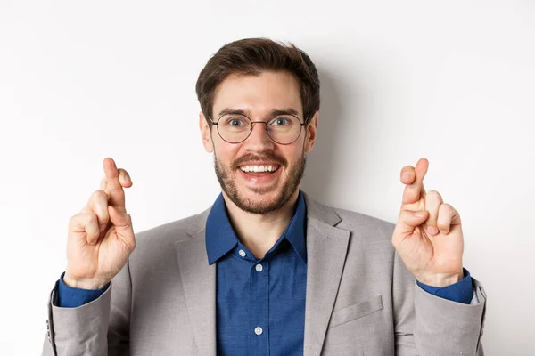 Primer plano del empresario masculino esperanzado en gafas y traje dedos cruzados para la buena suerte, sonriendo y pidiendo deseos, rezando por buenos resultados, fondo blanco — Foto de Stock