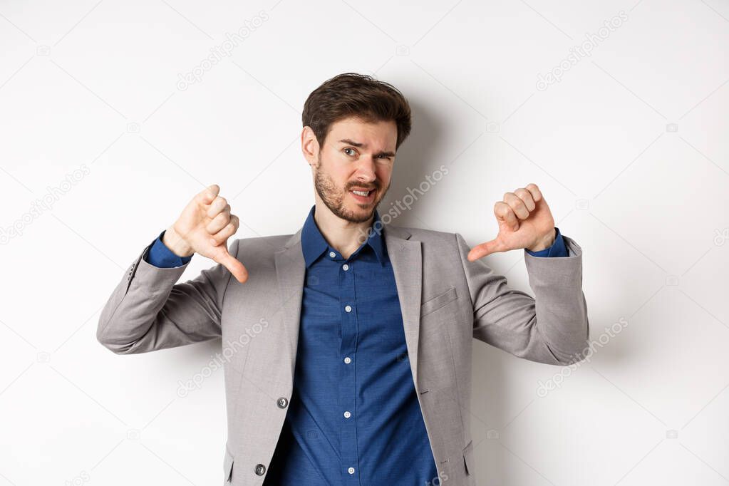 It sucks. Man feeling awkward showing thumbs down and judging something bad, frowning disappointed, standing in suit on white background