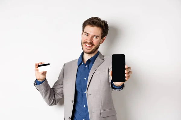 Online nakupování. Úspěšný podnikatel držící plastovou kreditní kartu a ukazující mobilní obrazovku, usměvavý spokojený, vydělávání peněz, bílé pozadí — Stock fotografie
