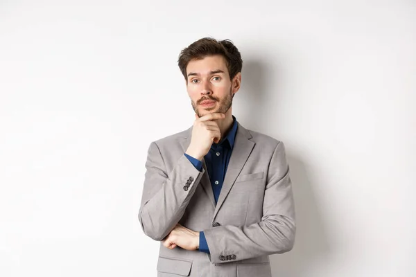 Pensativo hombre de negocios en traje tocando la barba, pensando y mirando a la cámara, decidiendo con cara pensativa, de pie sobre fondo blanco — Foto de Stock