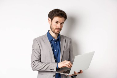 Takım elbiseli yakışıklı iş adamı dizüstü bilgisayarda çalışıyor, kameraya özgüvenli bakıyor, beyaz arka plana karşı duruyor.