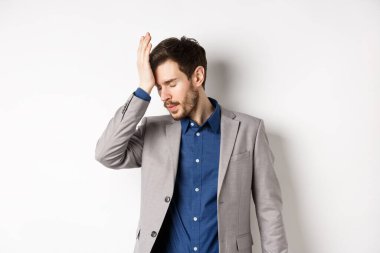 Annoyed businessman in suit facepalm with eyes closed, bothered by someone stupid, pissed-off at work, standing on white background