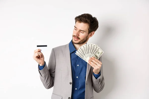 Homem de sucesso segurando notas de dólar e olhando para o cartão de crédito de plástico, satisfeito com o banco, de pé sobre fundo branco em terno de negócios — Fotografia de Stock