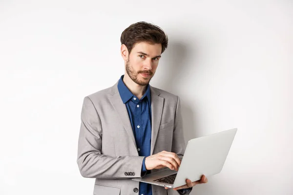 Knappe zakenman in pak werkend op laptop, kijkend naar camera zelfverzekerd, staande tegen witte achtergrond — Stockfoto