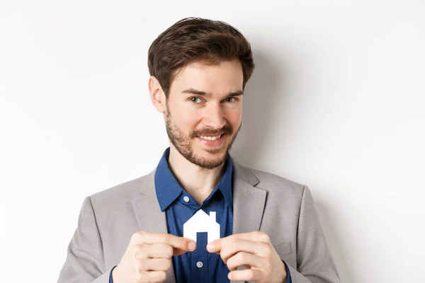 Concepto de bienes raíces y seguros. Sonriente agente inmobiliario masculino mostrando recorte de casa de papel pequeño y mirando a la cámara, la venta de la propiedad, fondo blanco — Foto de Stock