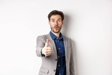 Takım elbiseli yakışıklı iş adamının görüntüsü. Heyecanlı yüzüyle baş parmağını gösteriyor. Beyaz arka planda duruyor.