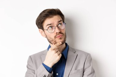 Sakallı ve gözlüklü düşünceli bir işadamı, karar veriyor, yukarı bakıyor ve düşünüyor, düşünceli yüzüyle takım elbise içinde duruyor, beyaz bir geçmişi var.