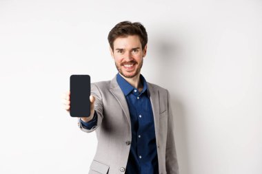 E-ticaret ve online alışveriş konsepti. Takım elbiseli kendine güvenen bir işadamı boş bir akıllı telefon ekranı ile elini uzatıyor, telefonla gösteriyor, beyaz arka plana karşı duruyor.