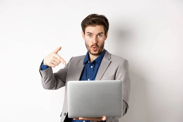 白い背景に対するビジネススーツに立って、驚くべき顔を持つノートパソコンの画面を指して、ここを見て求めて興奮ハンサムな男 — ストック写真