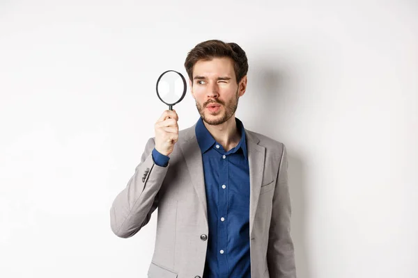 Snygg manlig modell i kostym tittar igenom förstoringsglas med intresse, ser något åt sidan, vit bakgrund — Stockfoto